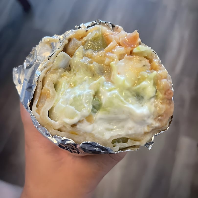 A hand holds a large, foil-wrapped El Faro Mexican Foods burrito filled with creamy sauce and ingredients.