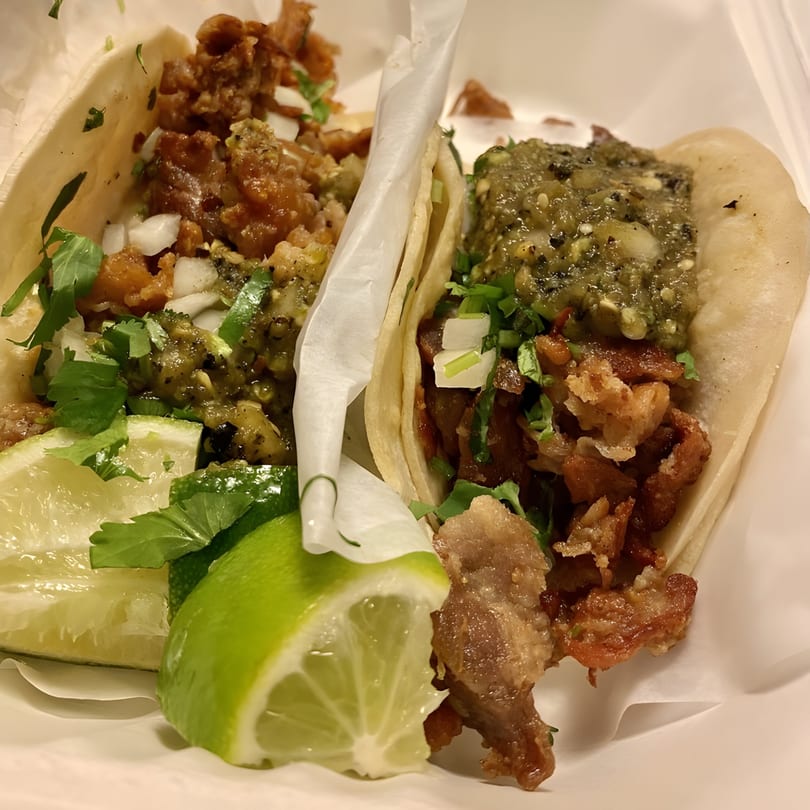 Two El Faro Mexican Foods tacos with meat, green salsa, cilantro, onions, and lime wedges.