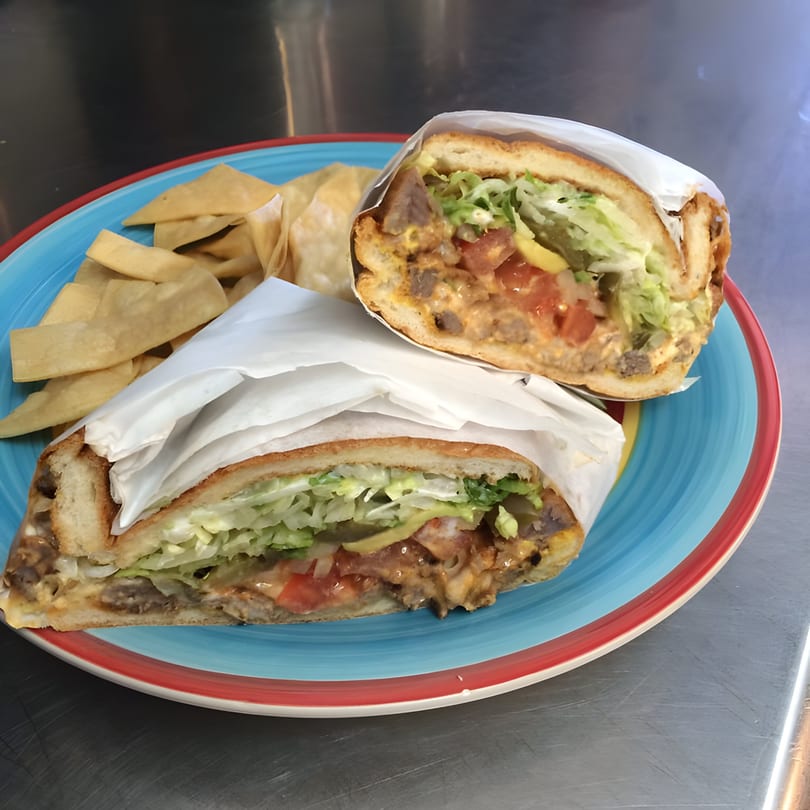 Two halves of a Mexican torta sandwich with meat, lettuce, and tomatoes, served with tortilla chips.
